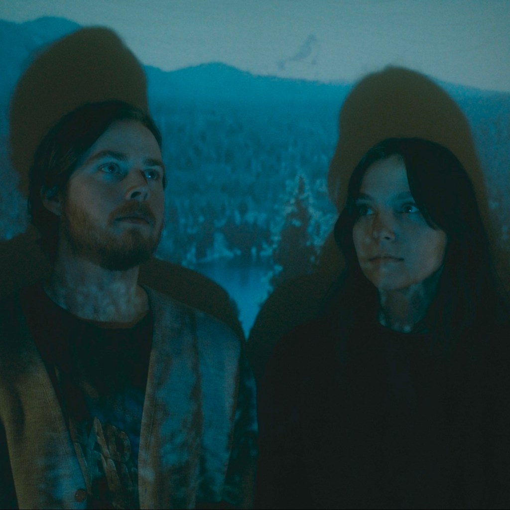 Band-mates Chris and Sam looking into the distance with a blue photo of a lake and mountains projected on to them.